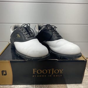 May include: A pair of black and white FootJoy golf shoes with black laces and silver eyelets. The shoes feature a textured leather upper, a white toe box, and a black saddle overlay. They are displayed on a black and gold FootJoy branded box.