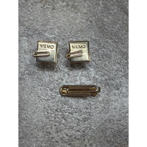 May include: Two gold-toned square charms, each featuring the word MEMO embossed on the surface with an attached gold-toned paperclip detail, accompanied by a single gold-toned paperclip-shaped accessory with a hinged clasp mechanism.