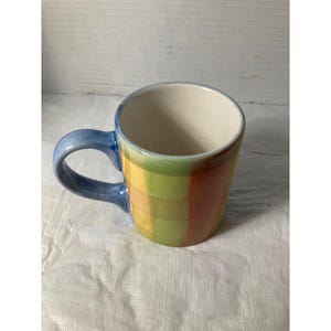 Pode incluir: Uma caneca de cerâmica com uma alça azul e um padrão xadrez colorido em tons de amarelo, verde e vermelho. O interior da caneca é branco. A caneca foi projetada para bebidas quentes.