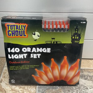 May include: A Totally Ghoul brand 140 orange light set in a rectangular box. The packaging features a haunted house, bats, and a moon graphic. The box indicates a total length of 12.8 metres, includes spare bulbs and a fuse, and is UL listed.