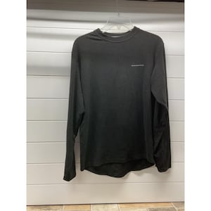 May include: A black long-sleeve shirt with a crew neckline and a small white text graphic on the upper left chest. The shirt features a straight hemline and is displayed on a clear plastic hanger against a white horizontal paneled wall.