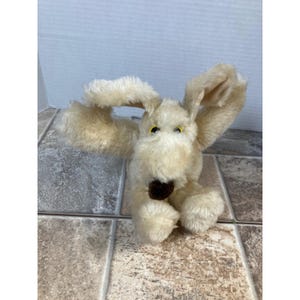 May include: A vintage plush dog toy with long, floppy ears and a brown nose. The dog has yellow eyes and is made of a soft, light brown material. The toy is sitting on a tiled surface.