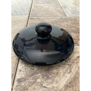May include: A circular, glossy black lid featuring a smooth, domed surface and a centered, rounded knob handle. The lid has a reflective finish and a simple, clean design, providing a secure cover for a container or pot with a matching circular opening.