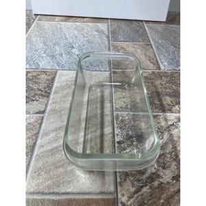 Puede incluir: Una fuente rectangular de vidrio transparente, con esquinas redondeadas y un asa en cada extremo. La fuente está vacía y se encuentra sobre una superficie de baldosas. El vidrio es transparente.