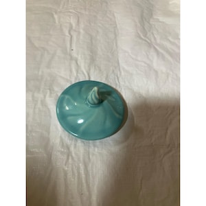 Pode incluir: Uma tampa redonda de cerâmica turquesa com um padrão de redemoinho e uma alça branca em espiral. A tampa é brilhante e parece ser para um frasco ou recipiente. O fundo é um tecido branco enrugado.