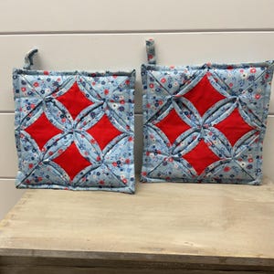 May include: Two square quilted potholders featuring a light blue fabric with a small floral pattern in red, white, and dark blue. Each potholder has a cathedral window design with four prominent red diamond-shaped centers and a fabric loop.