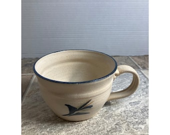 May include: A beige ceramic mug with a dark blue rim and handle. The mug features a dark blue abstract design on the side. The mug is sitting on a gray and brown tiled surface.