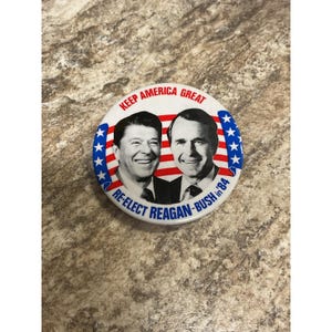 Peut inclure: Un bouton rond blanc avec le texte "KEEP AMERICA GREAT" et "RE-ELECT REAGAN-BUSH '84". Il présente des portraits de Ronald Reagan et George H.W. Bush, entourés d'étoiles et de rayures bleues.