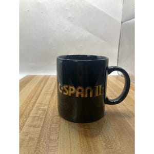 以下が含まれることがあります： 黒いセラミックマグカップ、湾曲したハンドル付き。 マグカップの前面には、金色の文字で「C-SPAN II」と書かれています。 マグカップは木製の表面に置かれています。