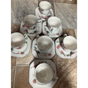 May include: A set of six white ceramic teacups and matching saucers, each featuring a delicate floral pattern with pink blossoms and green leaves. The saucers have an octagonal shape with thin dark green rims, and the cups have curved handles.