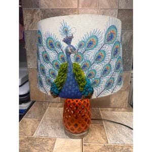 Peut inclure: Lampe de table décorative avec un motif de paon. L'abat-jour présente un paon avec des plumes bleues, turquoise et orange. Le socle est orange avec un motif en treillis.