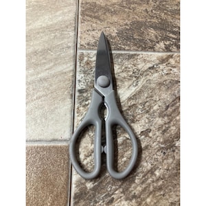 May include: A pair of gray handled scissors with silver blades. The scissors are open and resting on a patterned tile surface. The blades are sharp and pointed.