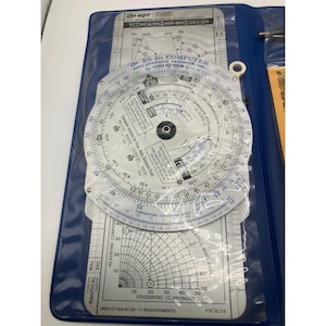 Könnte beinhalten: E6-B Flugcomputer für die Luftfahrt mit einem kreisförmigen Rechenschieber und gedruckten Skalen für Berechnungen. Der Computer befindet sich in einem blauen Vinyl-Etui mit einer transparenten Kunststoffabdeckung. Enthält eine Temperaturumrechnungsskala und eine Seitenwindkomponententabelle.