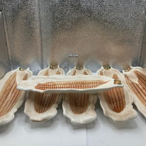 Może przedstawiać: Zestaw ceramicznych naczyń do serwowania w kształcie kolby kukurydzy. Naczynia są w kolorze ecru z brązowymi detalami imitującymi kukurydzę. Zestaw zawiera długi półmisek i kilka pojedynczych talerzy w kształcie kukurydzy.