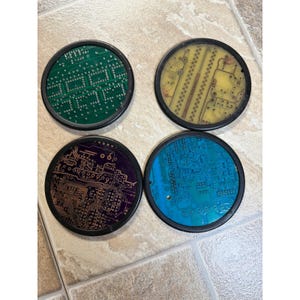 May include: Set of four round coasters with circuit board designs. Each coaster has a black border and features a different color and pattern: green, yellow, purple, and blue. The coasters are designed to protect surfaces from drink condensation.