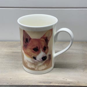 May include: A white ceramic mug featuring a detailed, realistic illustration of a brown and white Corgi dog on the front. The mug has a smooth, cylindrical body with a curved handle on the right side and a clean, minimalist white interior.