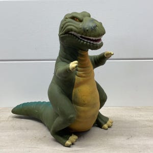 Könnte beinhalten: Ein geformtes Dinosaurier-Spielzeug aus Gummi mit einem dunkel olivgrünen Körper und einem kontrastierenden gelben Bauch. Die Figur hat eine texturierte Hautoberfläche, kleine Arme mit hellen Krallen und ein offenes Maul mit weißen Zähnen.