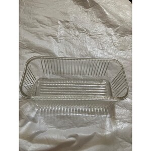 Puede incluir: Una fuente rectangular de vidrio transparente con detalles acanalados verticales y horizontales. La fuente tiene esquinas redondeadas y está vacía. La fuente está sobre una superficie blanca.