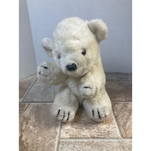 May include: A white plush polar bear toy with black eyes and a black nose. The bear is sitting upright with its arms outstretched. The bear's paws have black stitching to represent claws.