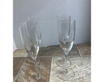May include: Four clear champagne flutes with long stems and round bases are arranged on a tiled surface. The glasses are empty and appear to be made of glass. The background is a plain white wall.