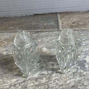 Può includere: Due saliere e pepiere in vetro trasparente e sfaccettato. Ogni shaker ha una parte superiore a cupola con piccoli fori e un corpo geometrico a forma di prisma. Gli shaker sono esposti su una superficie grigia maculata.