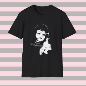 Peut inclure: Un t-shirt noir à manches courtes avec une impression graphique blanche représentant un visage de poupée et une figurine de poupée plus petite. Le texte "Nicole Dollanganger" est imprimé en police cursive sous le visage de la poupée.