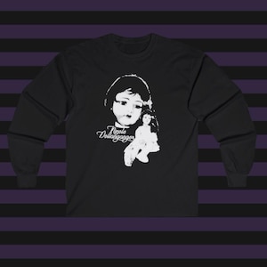 Peut inclure: Un t-shirt noir à manches longues avec une impression graphique blanche représentant le visage d'une poupée et une figurine de poupée plus petite. Le texte "Nicole Dollanganger" est imprimé en écriture cursive blanche sur le devant.