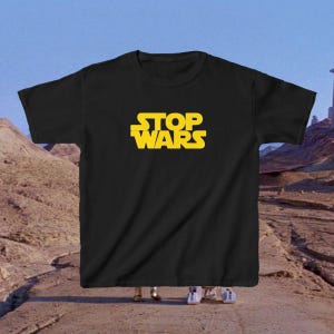 May include: A black short-sleeved t-shirt featuring the yellow text STOP WARS printed in a bold, stylized font across the center of the chest. The shirt has a classic crew neckline and a straight hem, presented against a desert landscape background.