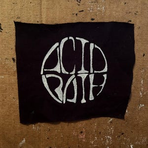 May include: A black fabric patch with the words "ACID BATH" in a white, stylized font, arranged in a circular design. The patch has a slightly frayed edge and is set against a brown, textured background.