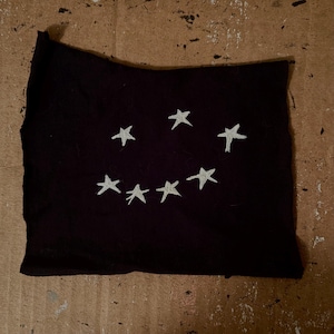 May include: A rectangular piece of black fabric featuring seven hand-embroidered white stars arranged in a curved, smiling pattern across the center. The fabric has raw, uneven edges and a soft, matte texture, showcasing a simple, stitched design.