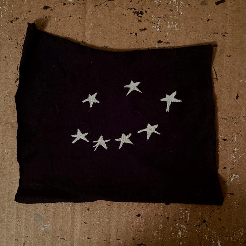 Peut inclure: Un morceau de tissu noir rectangulaire orn&eacute; de sept &eacute;toiles blanches brod&eacute;es &agrave; la main, dispos&eacute;es en arc de cercle pour former un sourire. Le tissu pr&eacute;sente des bords bruts et irr&eacute;guliers ainsi qu'une texture mate et douce au toucher.
