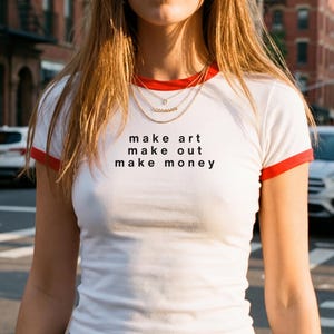 Op de afbeelding: Wit T-shirt met rode rand en de tekst "make art make out make money" in het zwart. De persoon draagt een blauwe spijkerbroek en een gouden ketting. De achtergrond toont een stadsstraat met auto's en gebouwen.