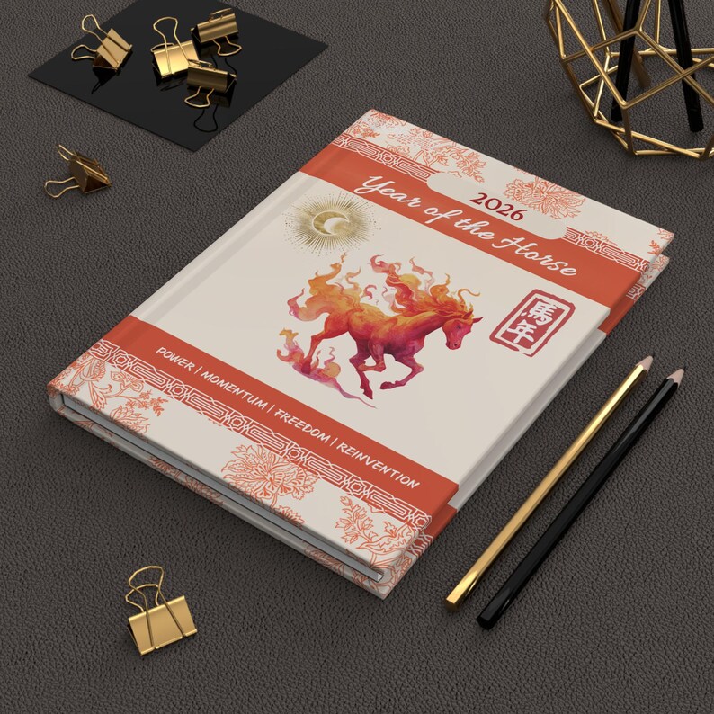 A hardcover journal with a cream-colored cover featuring a fiery horse illustration. The journal is labeled "Year of the Horse 2026" with decorative floral patterns and the words "POWER | MOMENTUM | FREEDOM | REINVENTION". Gold and black pencils and binder clips are nearby.
