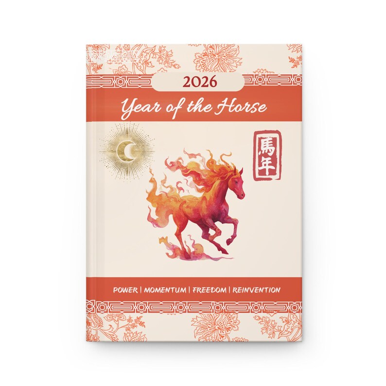 A hardcover journal with a cream-colored background and a watercolor illustration of a fiery horse. The cover reads "2026 Year of the Horse" in orange, with additional text: "POWER | MOMENTUM | FREEDOM | REINVENTION". Decorative floral patterns frame the text.