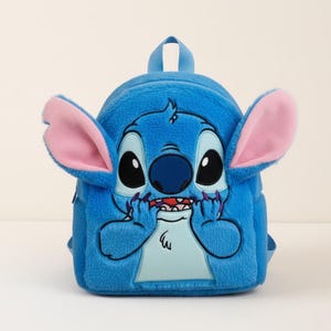 Puede incluir: Una mochila de felpa azul que representa al personaje Stitch de Lilo & Stitch. La mochila tiene orejas grandes con forro rosa, ojos grandes negros y una boca cosida. Tiene un asa superior peque&ntilde;a y correas ajustables.