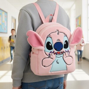 Puede incluir: Mochila de felpa rosa con un personaje de dibujos animados. La mochila tiene orejas rosas grandes, ojos grandes y una nariz azul. Cuenta con correas rosas y un peque&ntilde;o bolsillo delantero. La cara del personaje est&aacute; bordada con detalles negros y morados.