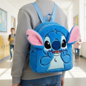 Puede incluir: Una mochila de felpa azul con el personaje Stitch de Lilo & Stitch. La mochila tiene grandes orejas rosas, una cara detallada y correas azules ajustables. La mochila la lleva una persona en un pasillo escolar.