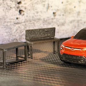 May include: A miniature scene featuring a red and black toy truck, a small table, and a bench. The truck has silver wheels and a black roof. The table and bench are dark grey. The background is a textured, light grey wall.