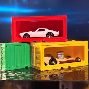 Puede incluir: Tres coches de juguete dentro de contenedores de envío en miniatura de colores. Un coche blanco está en un contenedor rojo, un coche beige está en un contenedor amarillo y un contenedor verde está vacío. Los coches están sobre una superficie reflectante.