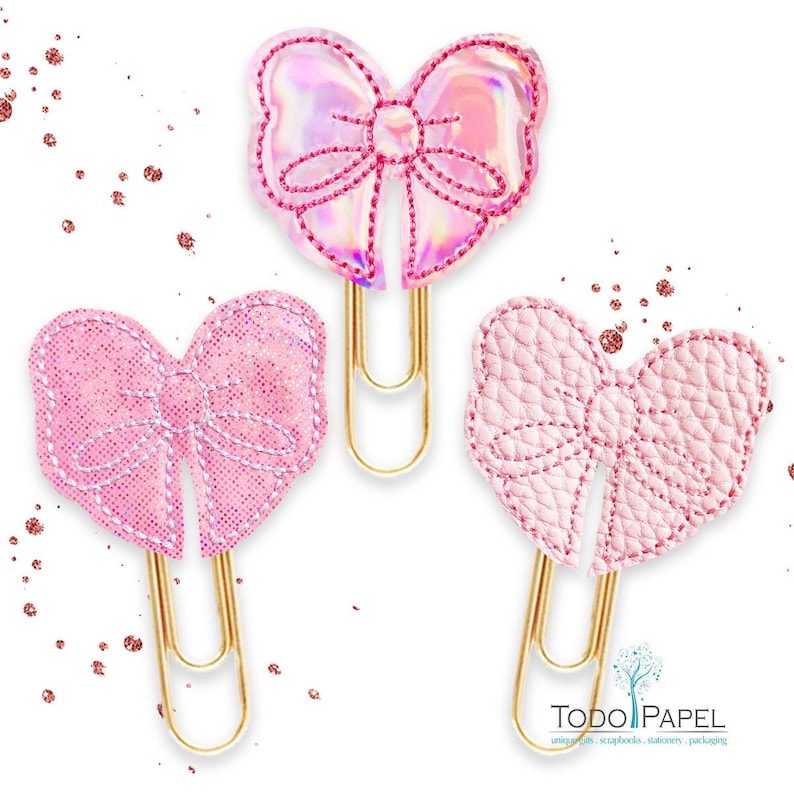 May include: Three gold paper clips with pink bow embellishments. The bows are made of different materials, including glitter, iridescent, and textured fabric.