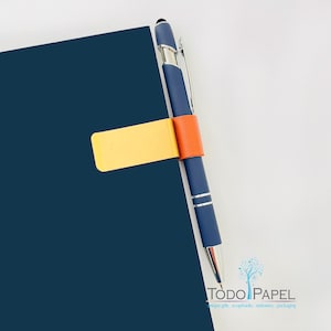 May include: A navy blue notebook with a yellow bookmark and an orange pen holder. A dark blue pen is held in place by the orange pen holder. The pen has silver accents. The notebook is on a white background.