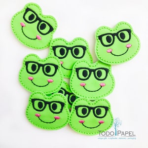 Könnte beinhalten: Sieben grüne Filz-Frosch-Applikationen mit schwarzen gestickten Brillen und rosa gestickten Wangen. Die Applikationen lächeln alle.