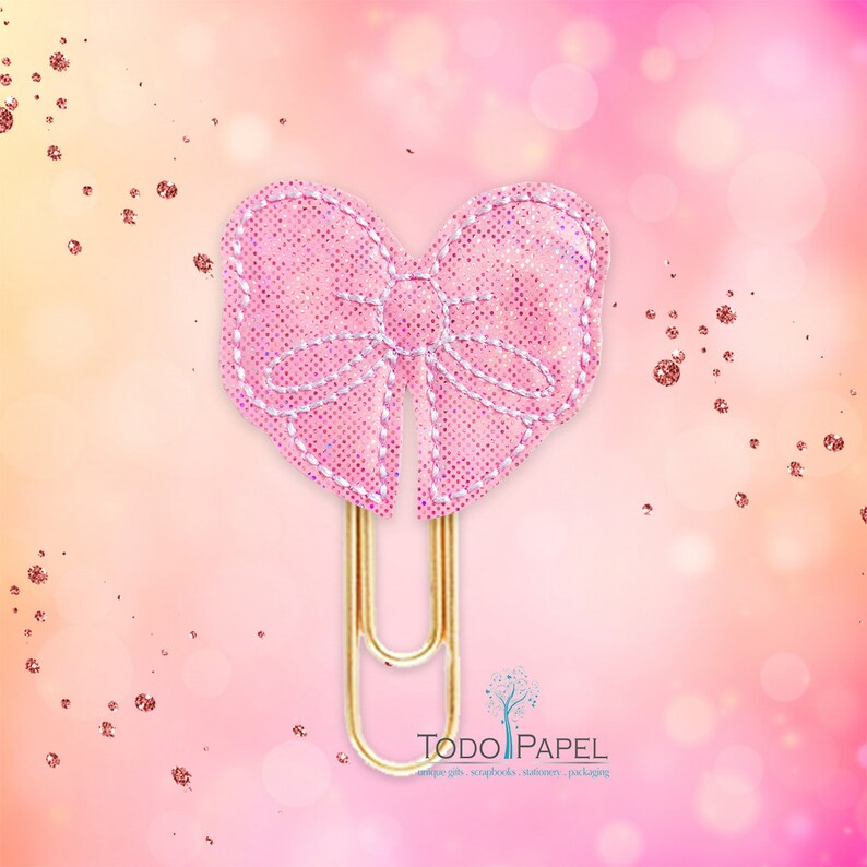 May include: A pink glitter bow paper clip with a gold metal clip. The bow is decorated with white stitching. The text "TODO PAPEL" is visible in the bottom right corner of the image.