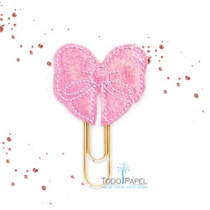 May include: A pink glitter bow paper clip with a gold metal clip. The bow is sewn with white thread and has a heart shape.