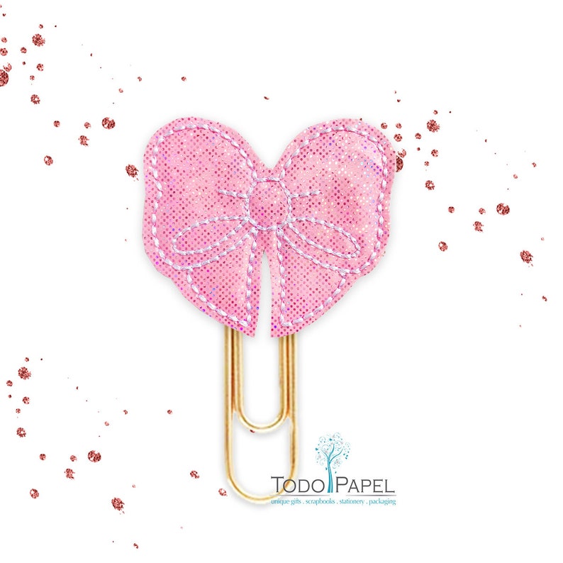May include: A pink glitter bow paper clip with a gold metal clip. The bow is sewn with white thread and has a heart shape.