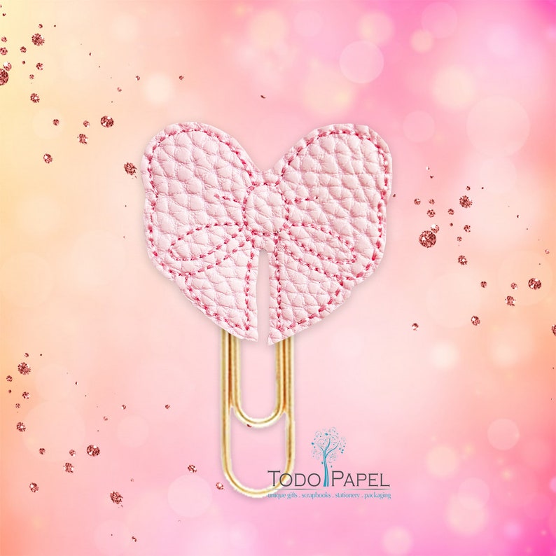 May include: A pink leather bow paper clip with gold metal accents. The bow is stitched with a pink thread. The paper clip is attached to the bow. The background is a pink and white gradient with gold glitter.