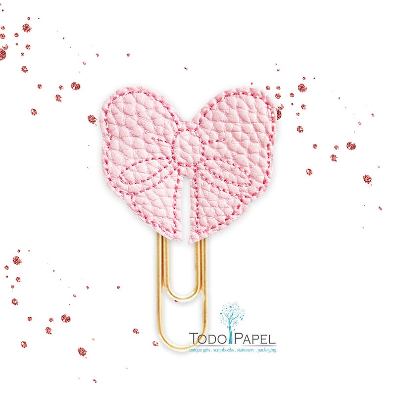 May include: A pink leather bow paper clip with a gold metal clip. The bow is stitched with a white thread.  "TODO PAPEL" is printed in black text on a white background below the clip.  "unique gifts, scrapbooks, stationery, packaging" is printed in black text below the logo.