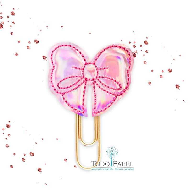 May include: A gold paper clip with a pink iridescent bow attached to the top. The bow has a pink stitched outline.