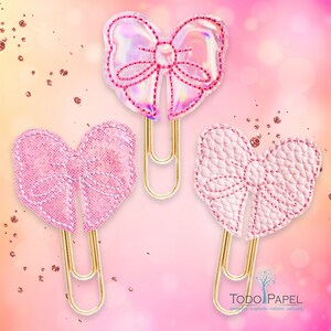 May include: Three gold paper clips with pink bows. The bows are made of different materials, including iridescent, glitter, and textured fabric.