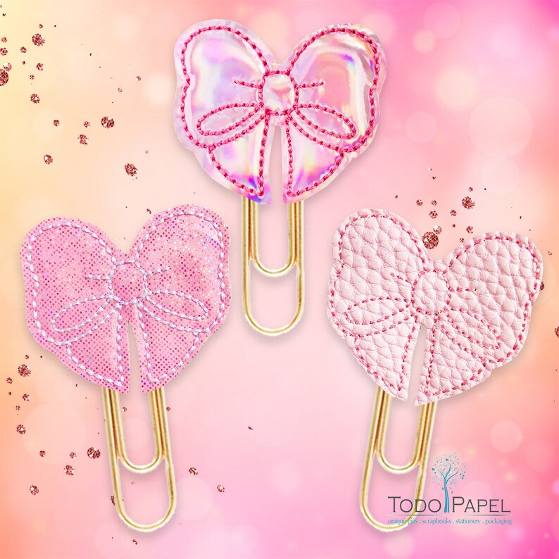 May include: Three gold paper clips with pink bows. The bows are made of different materials, including iridescent, glitter, and textured fabric.